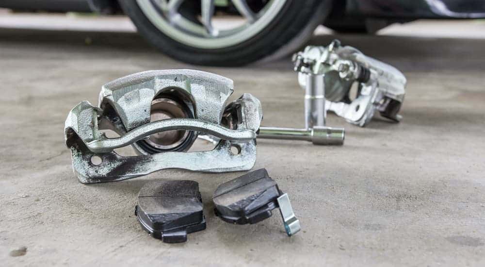 Calipers and brake pads are sitting on the concrete near a car.